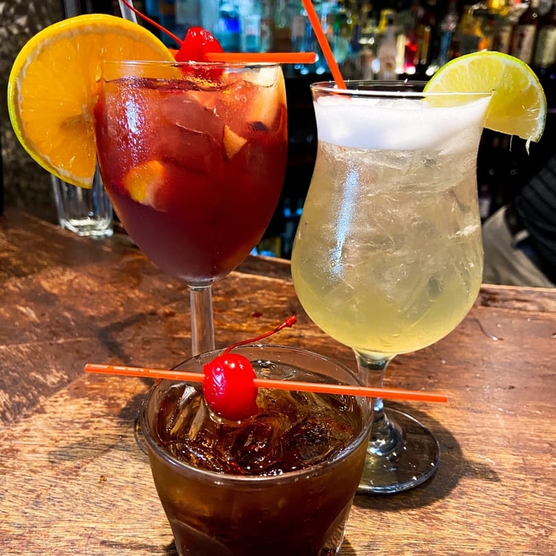 Three vibrant cocktails on a wooden bar, garnished with fruit and cherries, at Tre Scalini.