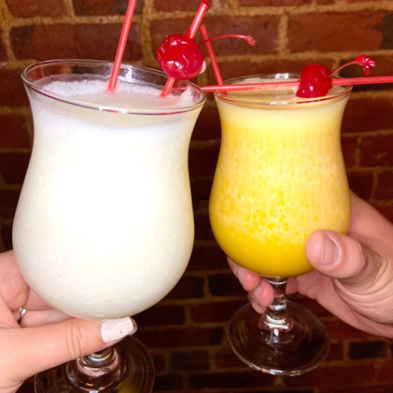Two hands holding up a white and a yellow frozen drink, garnished with cherries and straws.