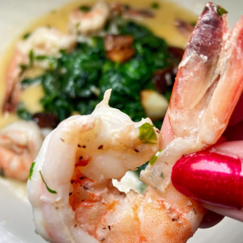 Close-up of a hand holding a shrimp, with a dish of shrimp and greens in the background.