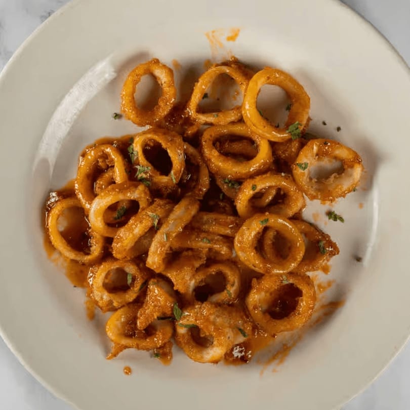 Plate of golden-brown fried calamari rings coated in a rich, reddish-orange sauce, garnished with herbs.