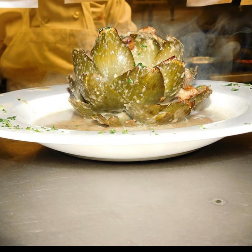 Steaming, whole baked artichoke in a white bowl with sauce, garnished with parsley.