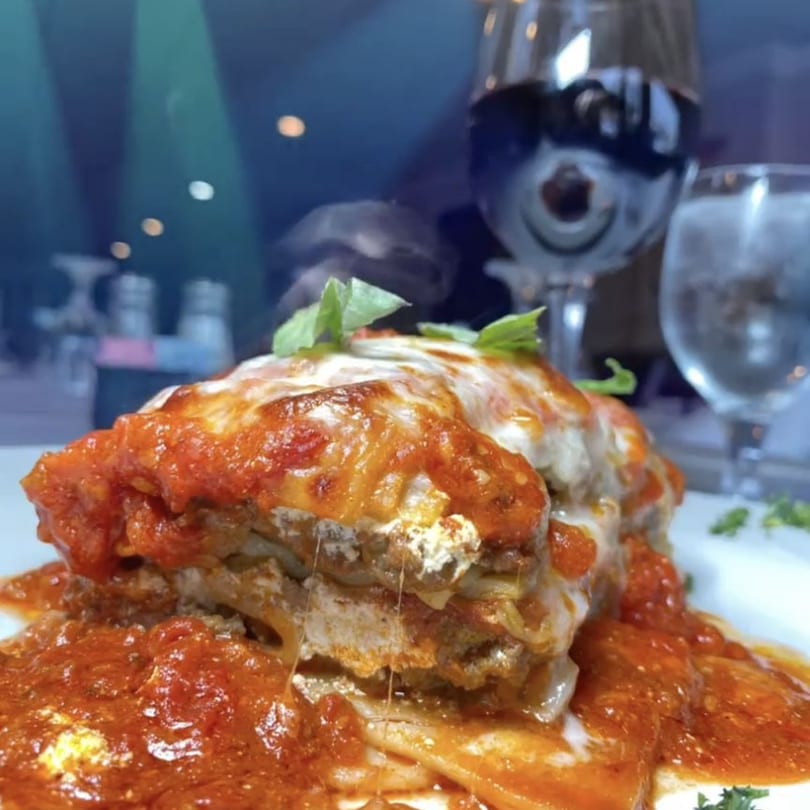 A steaming, cheesy lasagna with red sauce and basil, next to wine and water glasses.