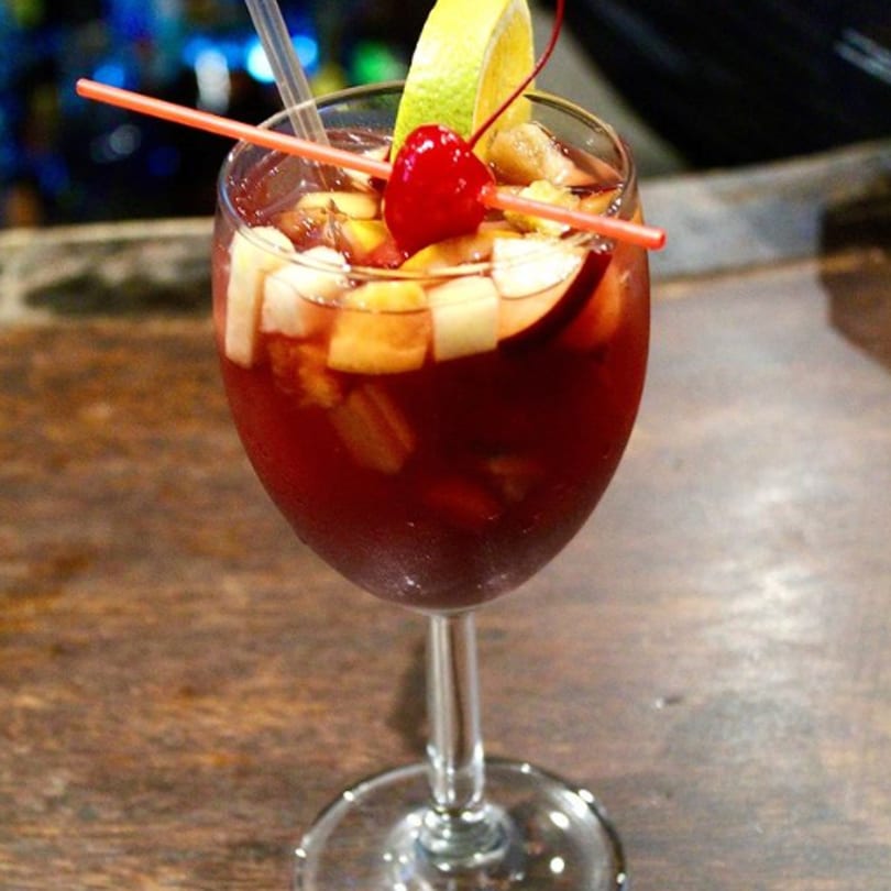 A glass of red sangria with fruit, a lime wedge, and a cherry on a wooden table.